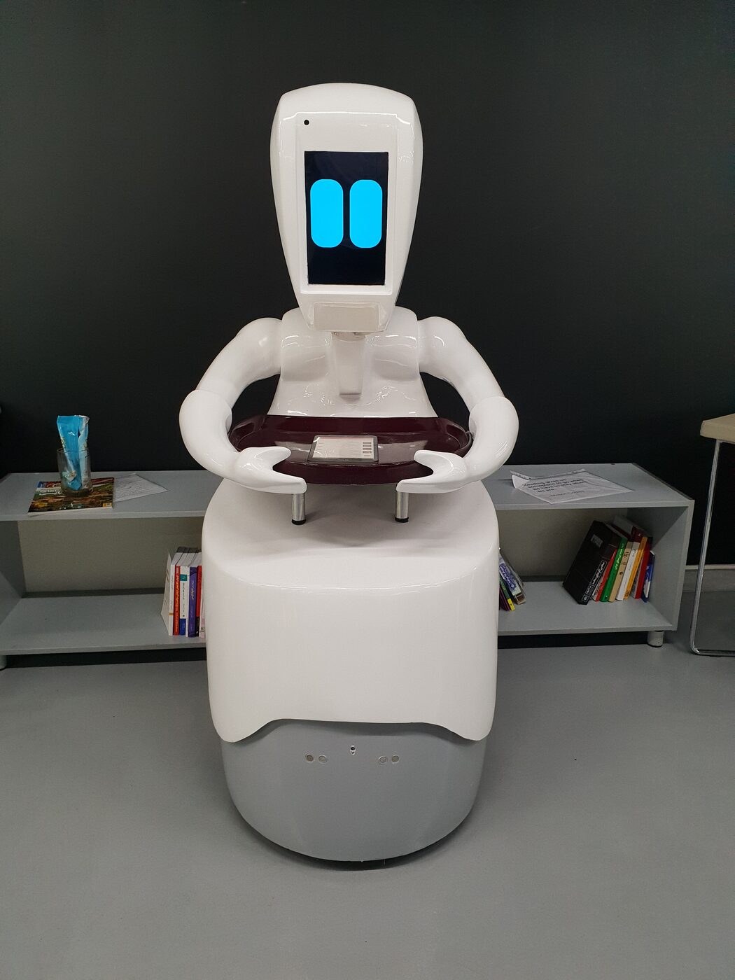 Hooshyar, a white service robot with a screen face and two arms supporting a tray, standing in an office lobby