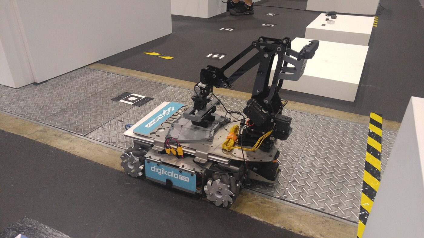 Mecanum-wheeled AGV carrying a multi-jointed manipulator arm, on a competition floor marked with fiducial tags